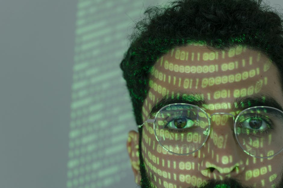 Close-up of a young man with curly dark hair and round glasses, facing forward with a focused expression. Overlaid on his face and glasses are green digital binary code and data streams, creating a vi