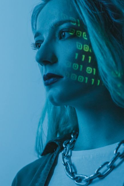 Close-up of a young man with curly dark hair and round glasses, facing forward with a focused expression. Overlaid on his face and glasses are green digital binary code and data streams, creating a vi