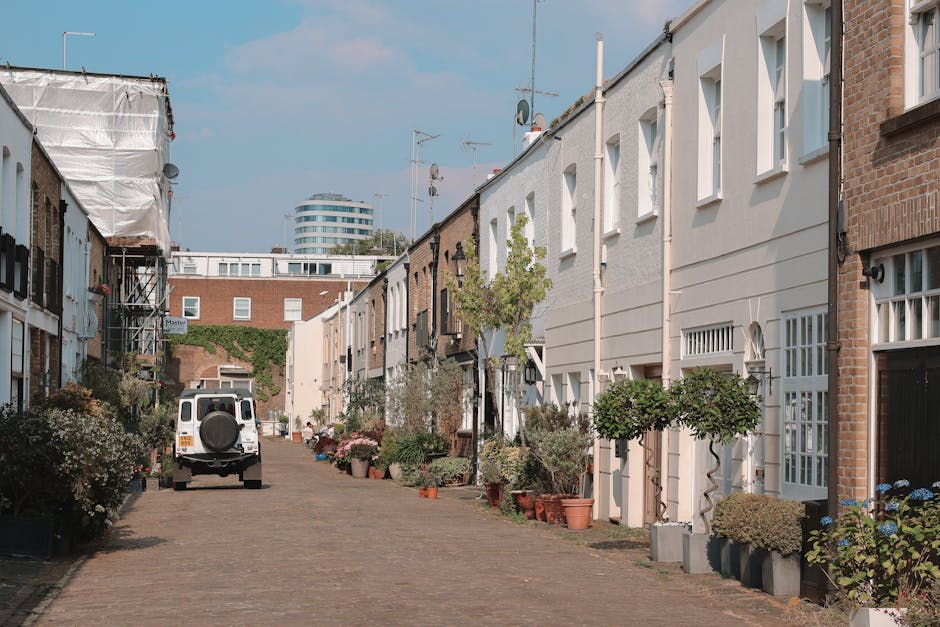 A quiet residential street lined with modern terraced houses featuring white and brick exteriors, decorative window frames, and small front gardens filled with potted plants and trimmed shrubs. The co