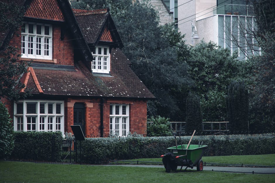 A quiet residential street lined with modern terraced houses featuring white and brick exteriors, decorative window frames, and small front gardens filled with potted plants and trimmed shrubs. The co